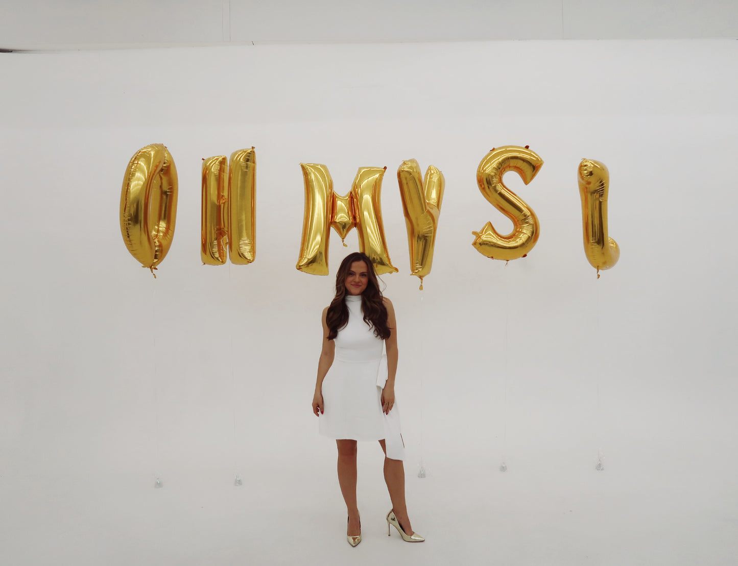 Woman in a white dress standing in front of gold letter balloons spelling 'OH MY SI' on a white background