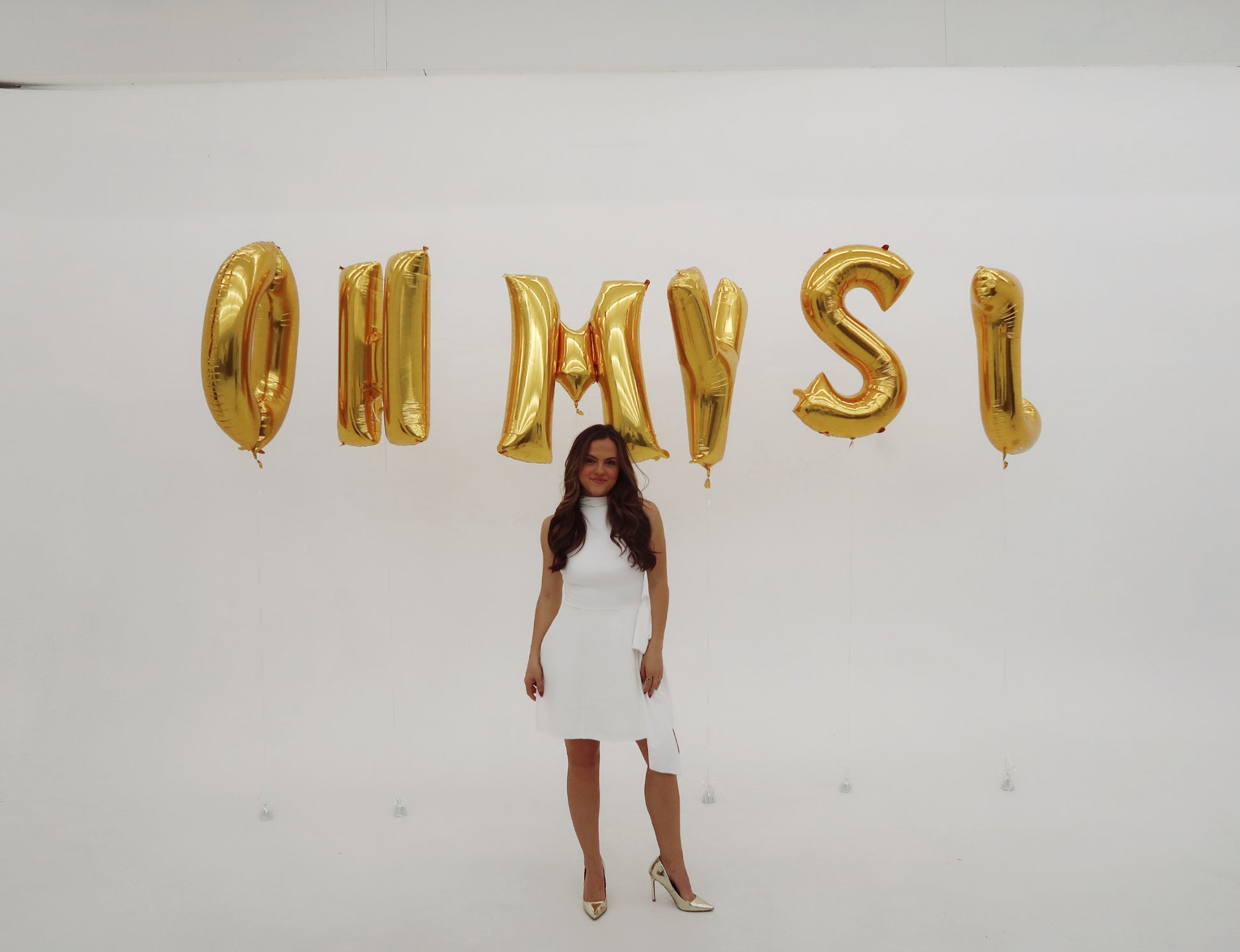 Woman in a white dress standing in front of gold letter balloons spelling 'OH MY SI' on a white background